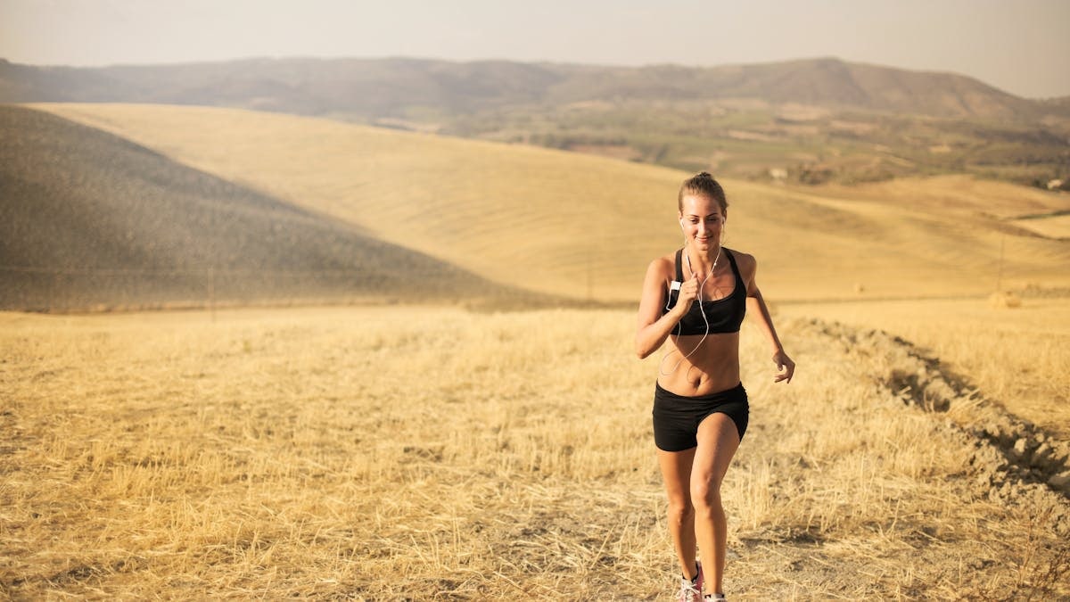 Powerful female athlete in sportswear and earphones jogging through field in sunny hot day